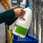 Mid-adult woman shopping on a budget. She is looking for low prices due to inflation, standing and holding a milk carton. She is living in the North East of England.
