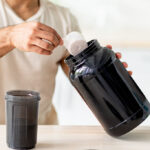 Unrecognizable young sporty guy making protein shake at table in kitchen, closeup. Cropped view of millennial bodybuilder using sports nutrition supplement, preparing cocktail indoors